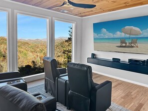 Ein modernes Wohnzimmer mit großem Fenster und Blick auf eine Berglandschaft, einem wandmontierten Flachbildfernseher und einer Sitzecke mit Ledersesseln.