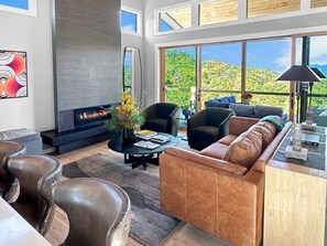 A modern living room with a fireplace, leather furniture, and large windows offering a view of greenery. - Mountain Modern 3BR cabin with hot tub, fire pit, lake access free train and rafting (Bryson City)