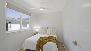 A bedroom with a bed, a ceiling fan, a window with blinds, and a nightstand with a lamp.
