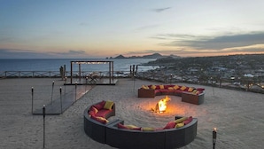 Un espace aménagé près de la plage, avec un foyer, du mobilier d’extérieur et une vue sur l’océan et les montagnes lointaines.