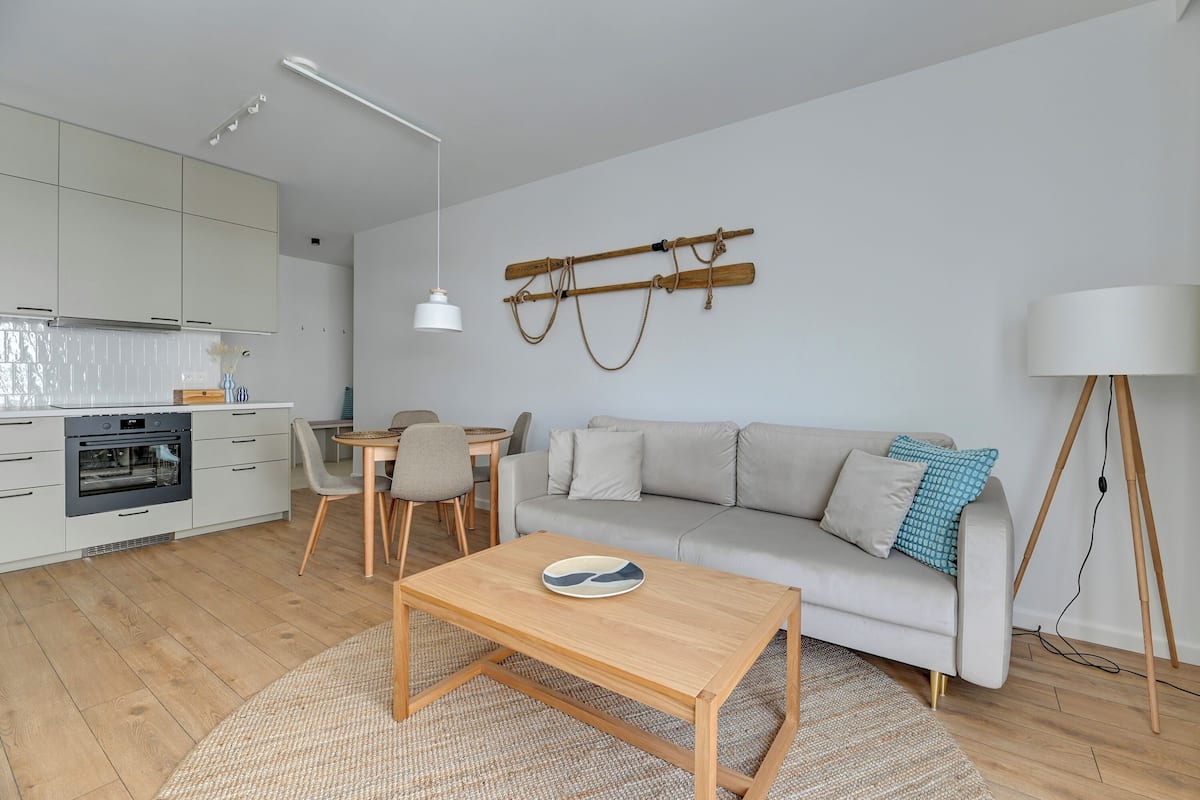 Eine moderne Küche mit weißen Schränken, ein Essbereich mit Holztisch und Stühlen, eine Couch mit Kissen und eine Stehlampe.