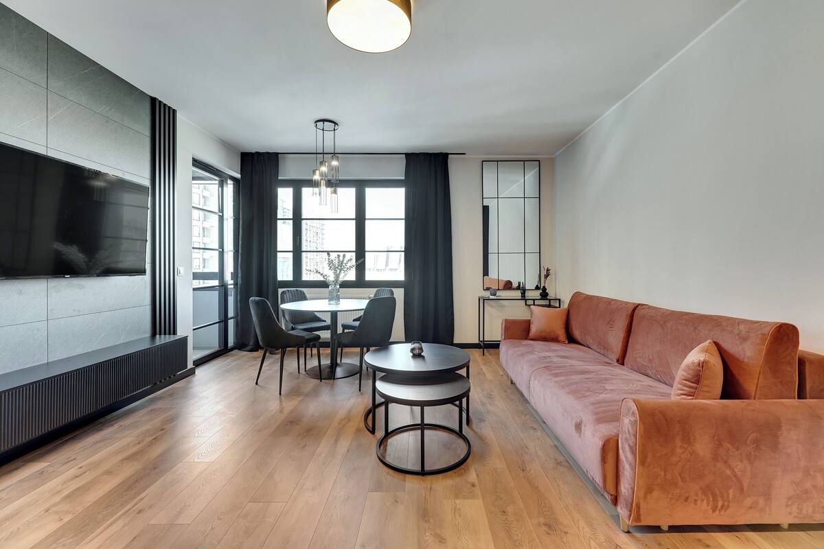 Ein modernes Wohnzimmer mit einer Couch, einem Esstisch mit Stühlen, einem Fernseher und einem großen Fenster mit Vorhängen.