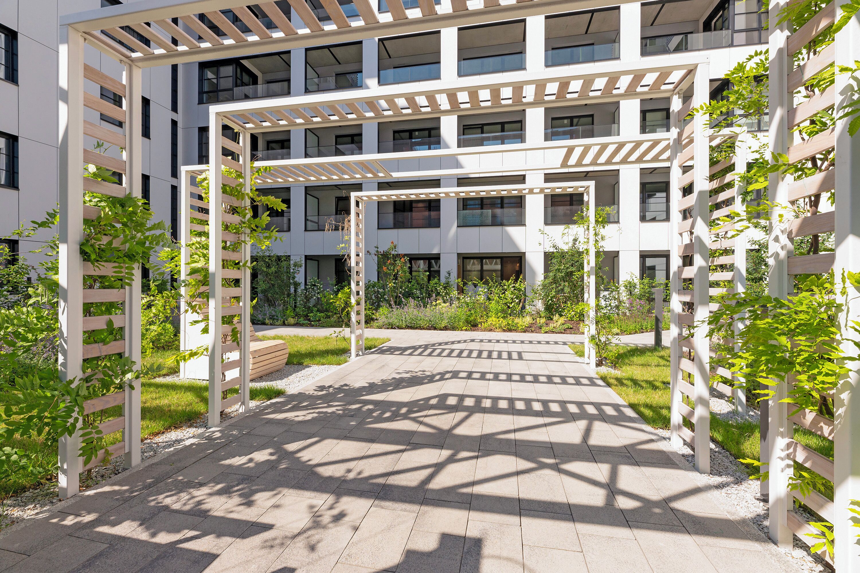 Un edificio moderno con varios pisos, un espacio exterior cubierto con una pérgola y zonas verdes alrededor.