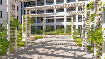 Un edificio moderno con varios pisos, un espacio exterior cubierto con una pérgola y zonas verdes alrededor.