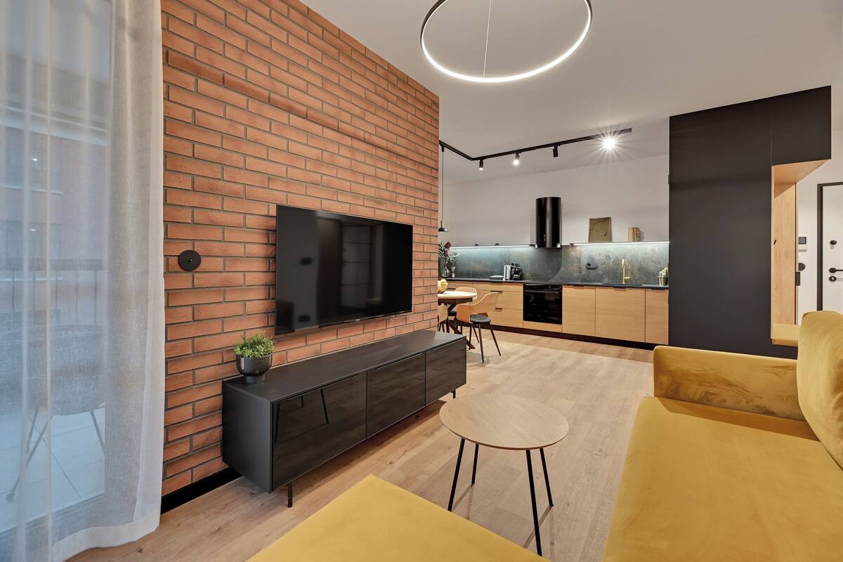 Ein modernes Wohnzimmer mit Backsteinwand, Flachbildfernseher, gelbem Sofa und einem kleinen runden Tisch.