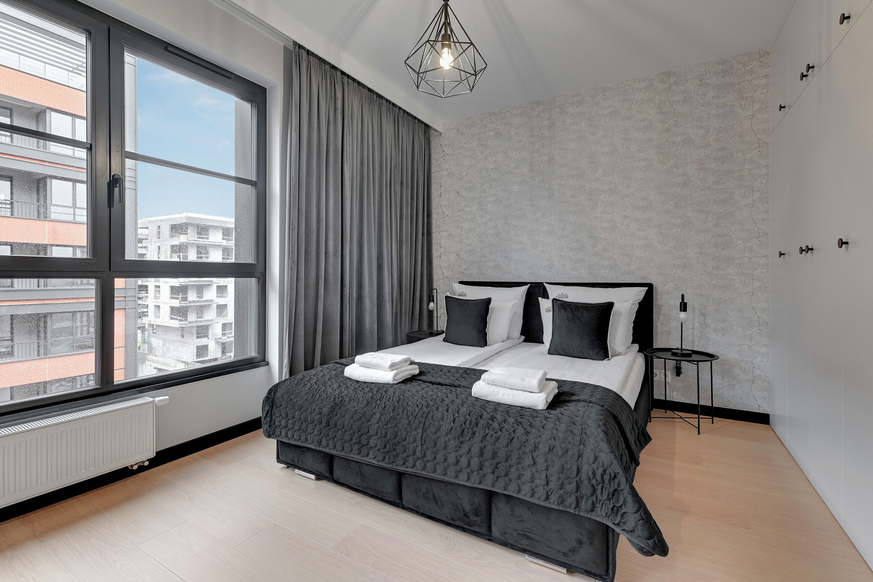 Een moderne slaapkamer met een groot raam, een bed met zwart-wit beddengoed, een klein nachtkastje en een radiator.