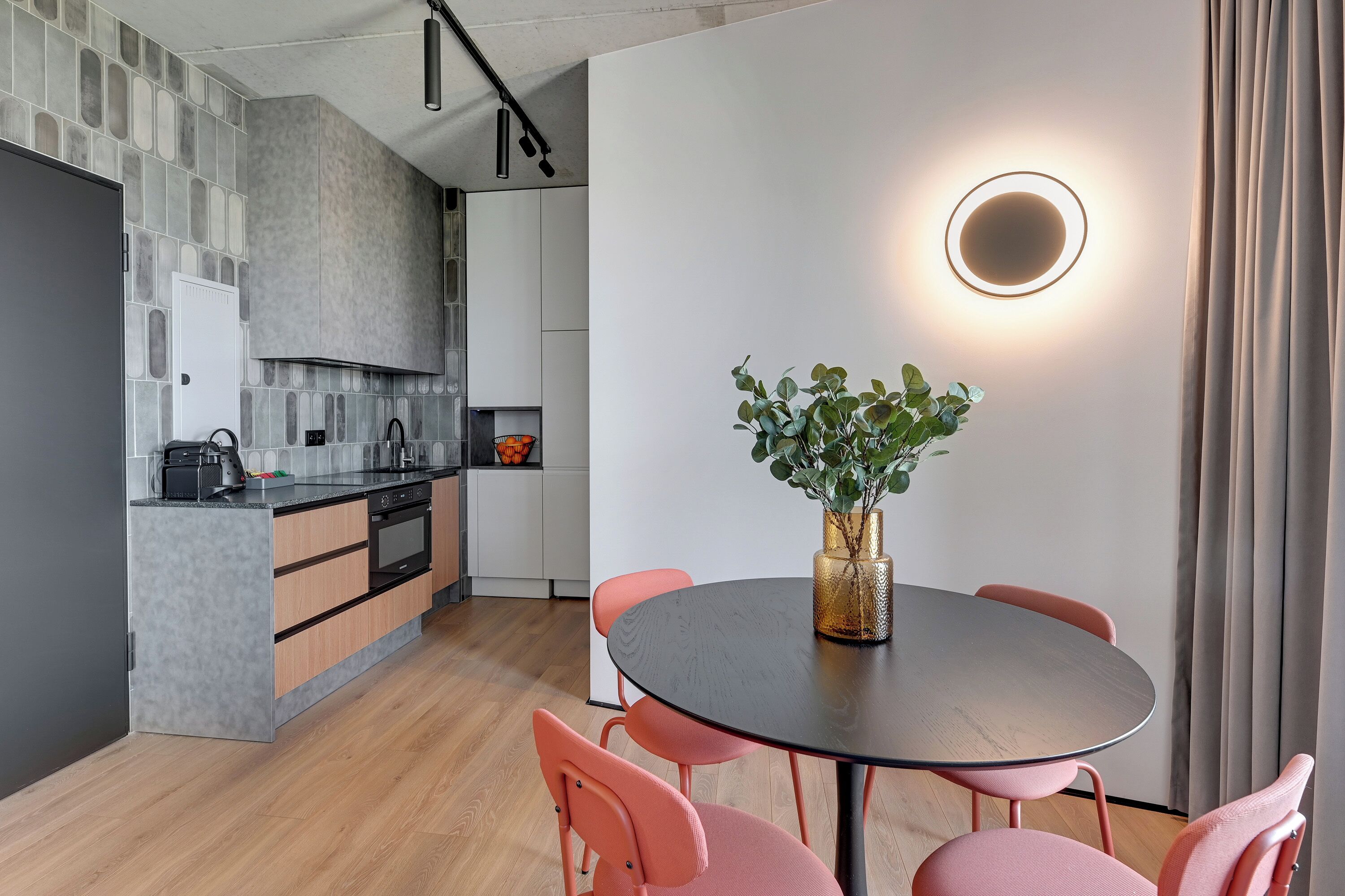 A modern kitchen with grey tiles, wooden cabinets, and a black countertop. There is a dining area with a round table and pink chairs, and a vase with green plants.