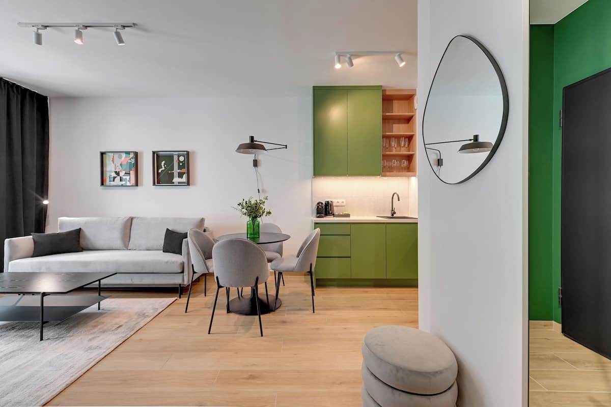 Ein modernes Wohnzimmer mit Sofa, Esstisch, Stühlen und Couchtisch. Es gibt eine Küchenzeile mit grünen Schränken und einer Spüle. Ein runder Spiegel hängt an der Wand.