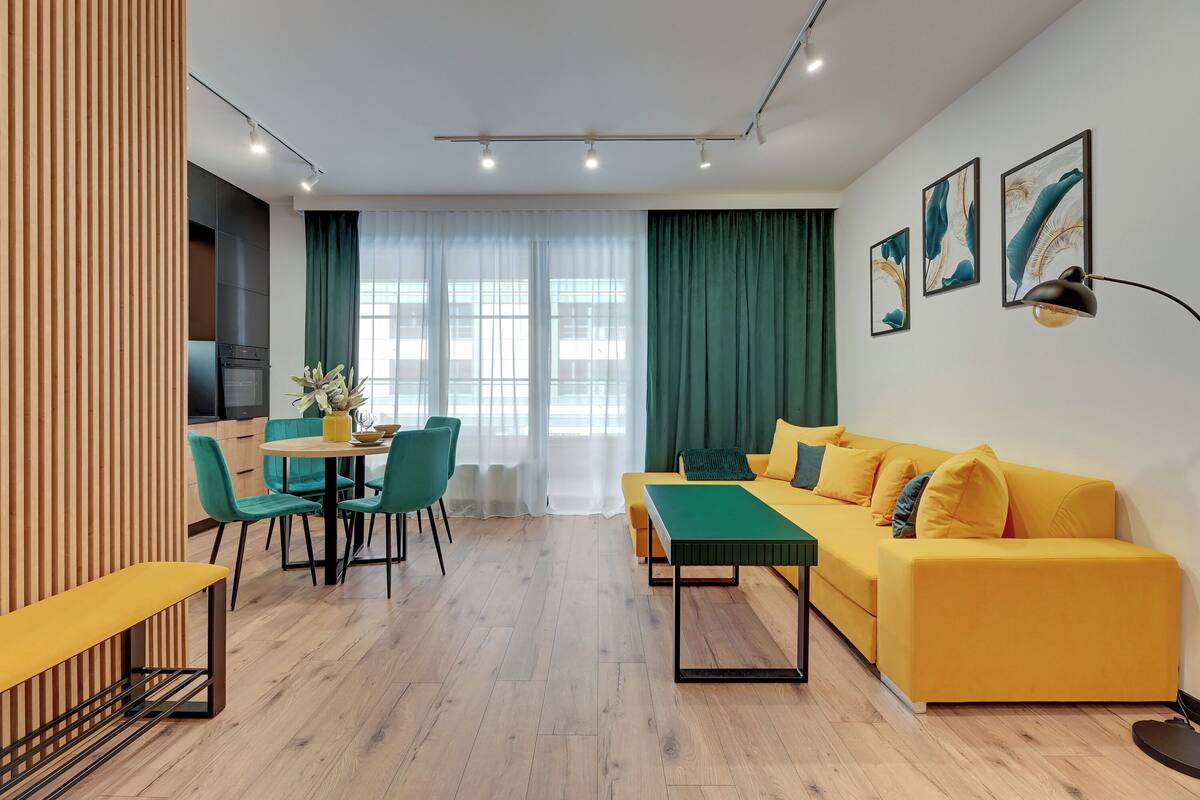 Ein modernes Wohnzimmer mit einem gelben Sofa, einer grünen Bank und einem Essbereich mit türkisfarbenen Stühlen und einem Holztisch.