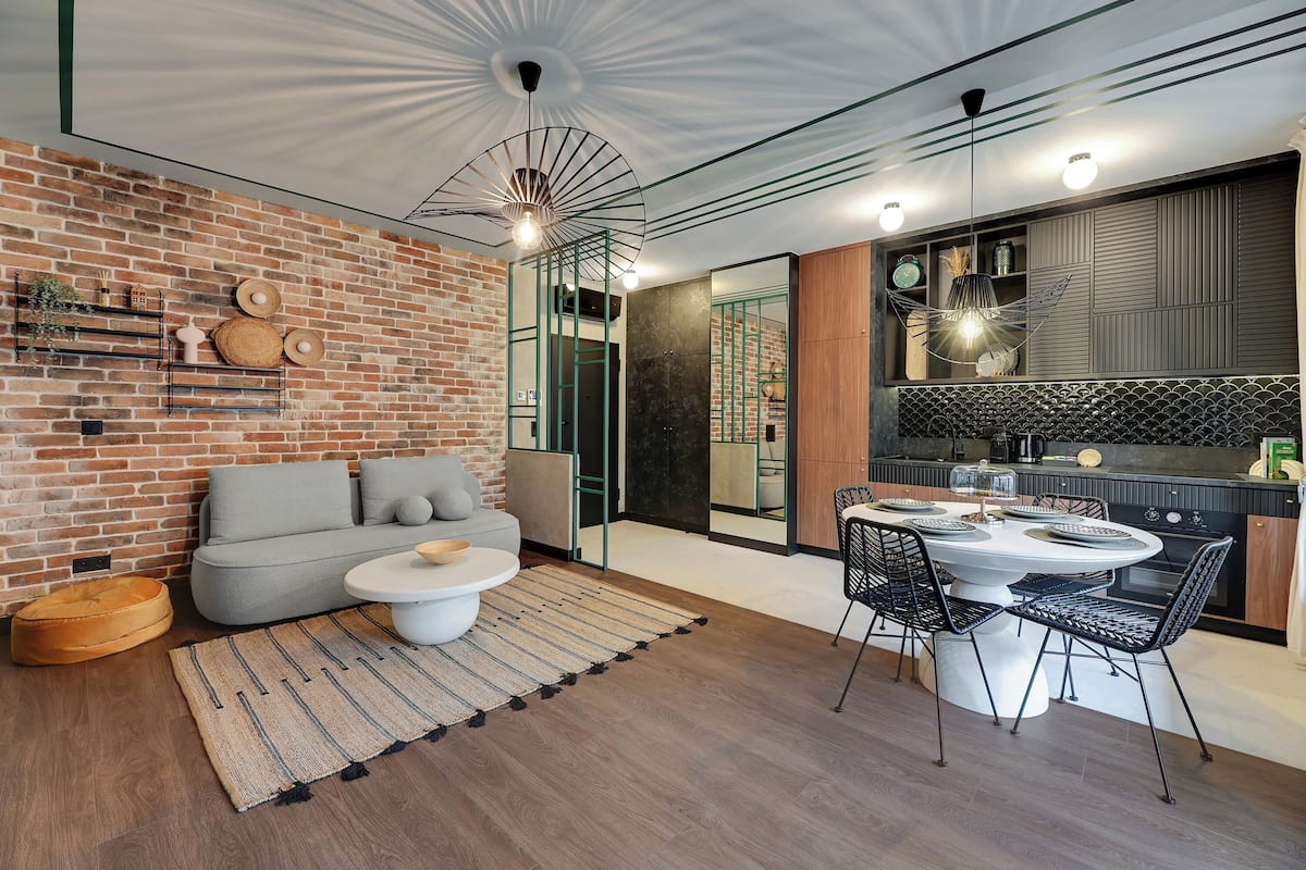 Ein modernes Wohnzimmer mit Backsteinwand, grauem Sofa, rundem Couchtisch und einem Essbereich mit weißem Tisch und schwarzen Stühlen.