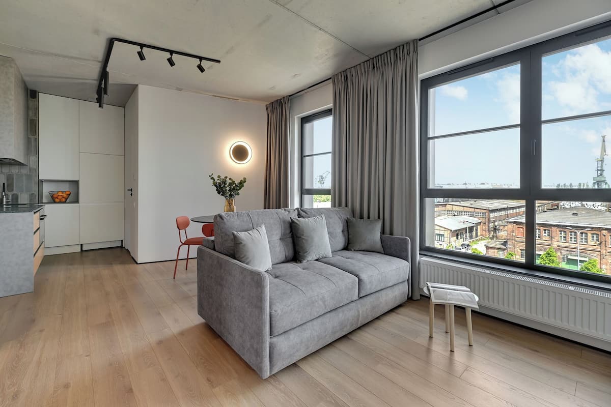 Ein modernes Wohnzimmer mit grauem Sofa, einem kleinen Tisch und Blick auf die Stadt durch große Fenster.