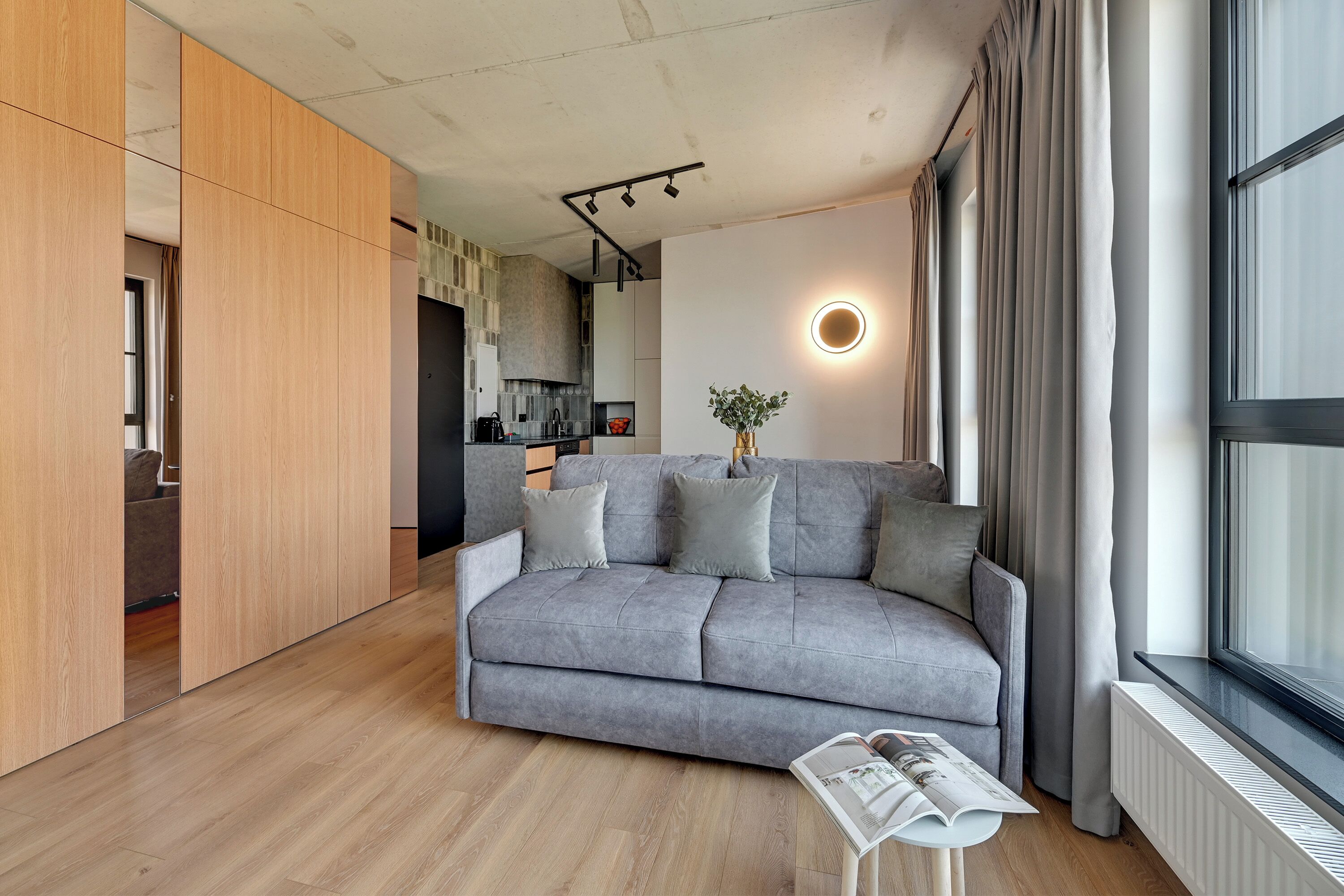 Ein modernes Wohnzimmer mit grauem Sofa, Holzboden, einer runden Wandleuchte und einem Fenster mit Vorhängen.
