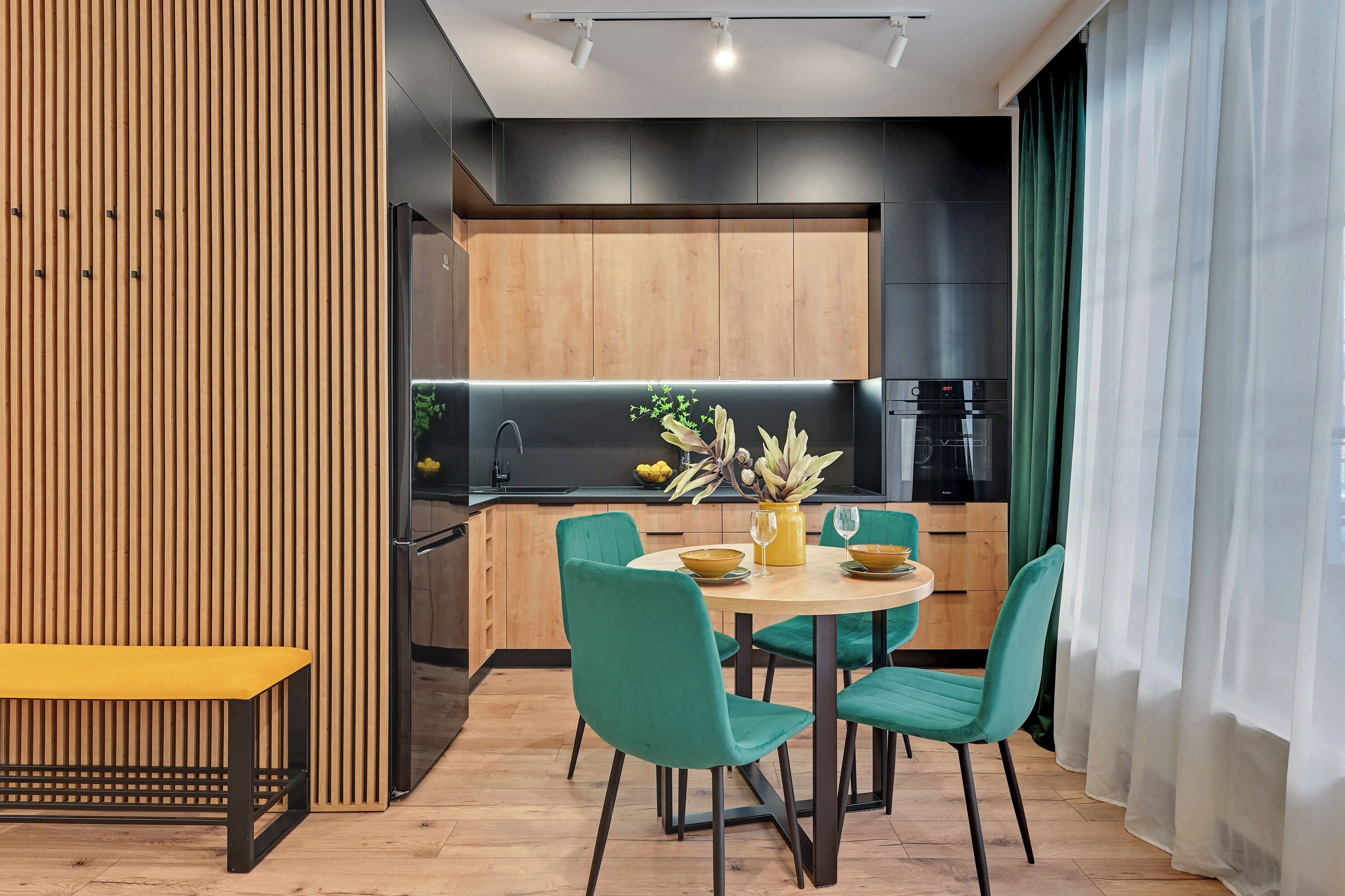 A modern kitchen with wooden cabinets, a round dining table with green chairs, and a yellow bench.