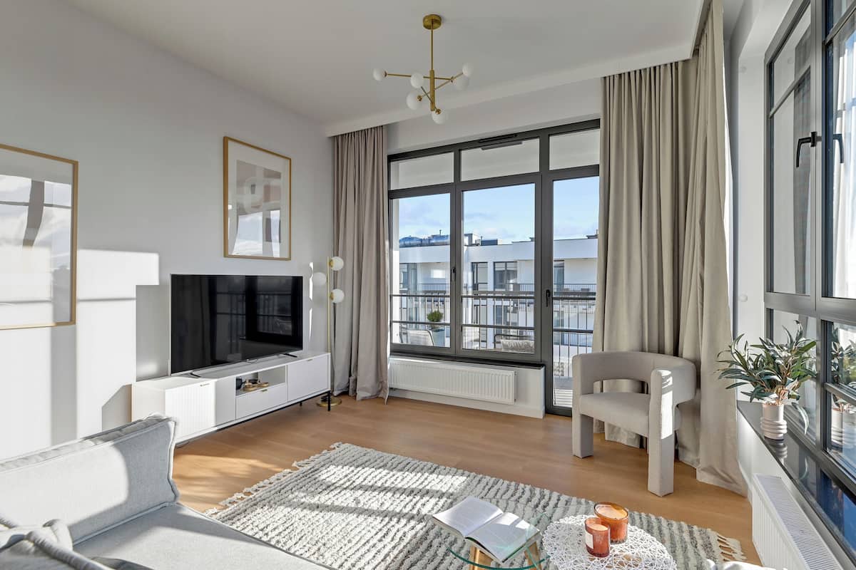 Ein modernes Wohnzimmer mit großem Fenster, einem Flachbildfernseher, einer Couch, einem Sessel und einem kleinen Tisch mit Buch und Kerzen.