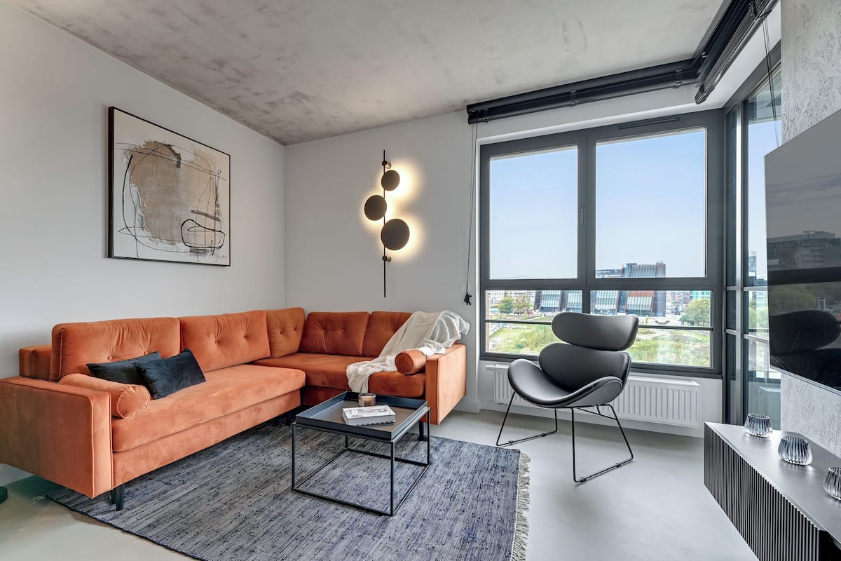 Ein modernes Wohnzimmer mit einem Ecksofa, einem Couchtisch, einem Sessel und einem großen Fenster mit Blick auf die Stadt.