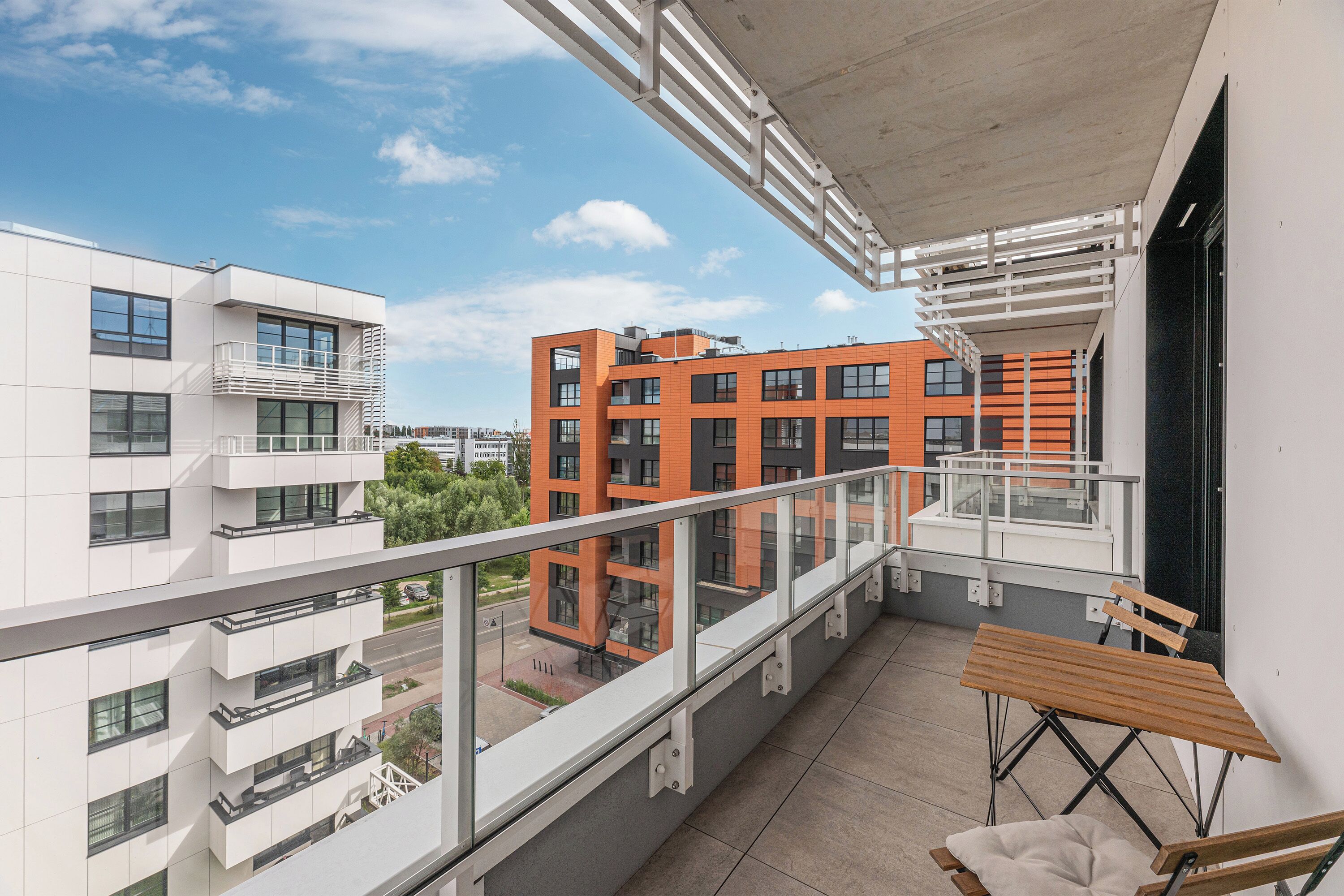 Un balcón con mesa y sillas de madera, con vistas a un edificio moderno de fachadas anaranjadas y blancas.