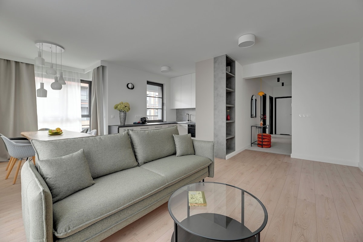 Ein modernes Wohnzimmer mit grauem Sofa, einem Glastisch und einer Küche im Hintergrund.