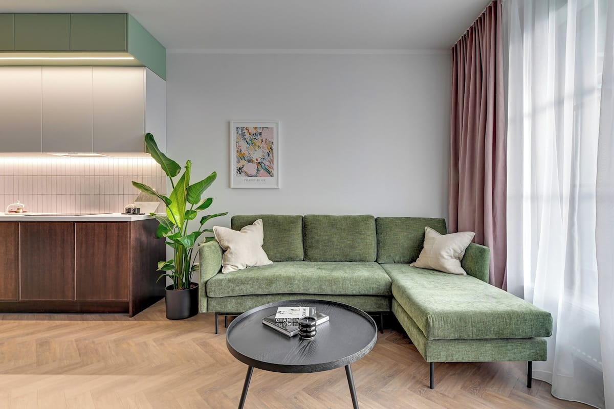 Ein modernes Wohnzimmer mit einem grünen Ecksofa, einem runden schwarzen Couchtisch, einer Topfpflanze und einer Küchenzeile mit weißer Arbeitsplatte und grünen Schränken.