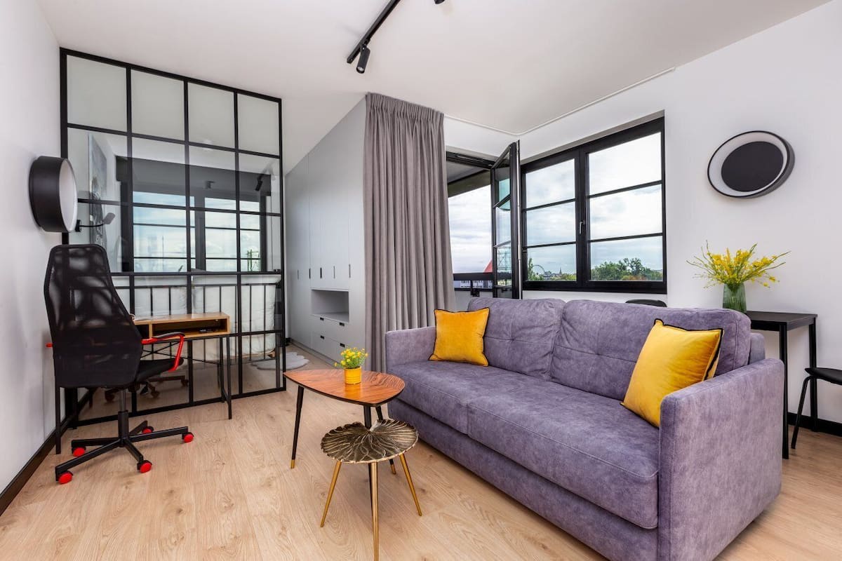 Ein modernes Wohnzimmer mit einem violetten Sofa, einem schwarzen Bürostuhl, einem Holztisch und Blick auf die Stadt durch große Fenster.