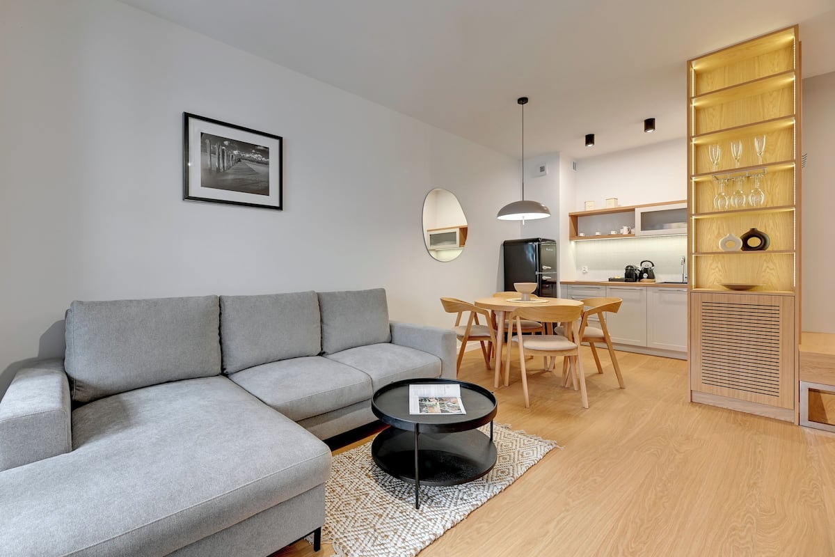 Ein modernes Wohnzimmer mit einer grauen Couch, einem runden Couchtisch und einem Essbereich mit Holztischen und -stühlen.