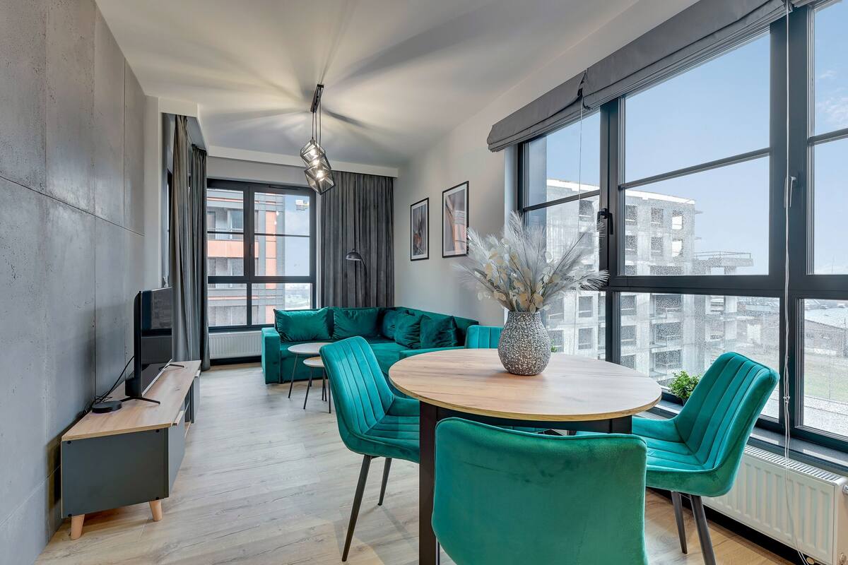Ein modernes Wohnzimmer mit einem türkisfarbenen Sofa, einem runden Holztisch und einem großen Fenster mit Blick auf die Stadt.