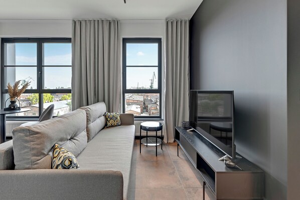 A modern living room with a sofa, a small round table, a flat-screen TV, and large windows with curtains. - Doki Living - Foodhall & Parking by Downtown Apartments (Gdansk)