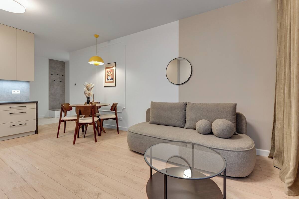 Ein modernes Wohnzimmer mit einer Couch, einem Glastisch, einem Essbereich mit Stühlen, einer Pendelleuchte und einem runden Spiegel an der Wand.