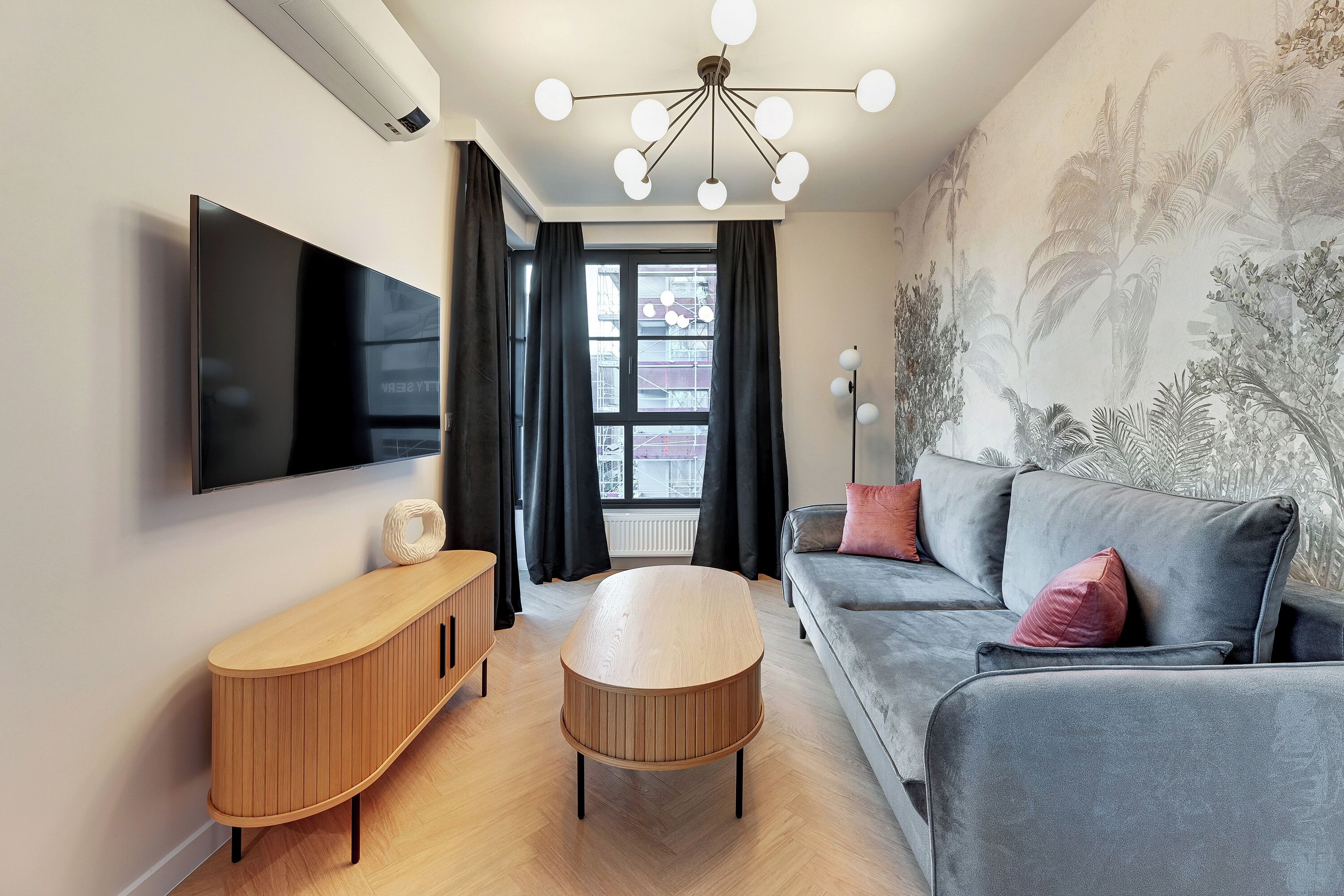 Ein modernes Wohnzimmer mit Flachbildfernseher, einem Holzkonsolentisch, einem runden Couchtisch, einer grauen Couch mit roten Kissen und einem großen Fenster mit schwarzen Vorhängen.