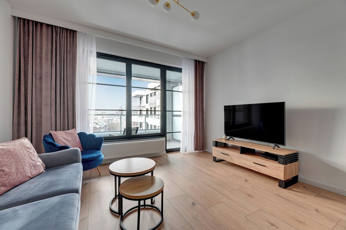 Een moderne woonkamer met een bank, twee ronde tafels, een blauwe fauteuil, een televisie en een groot raam met gordijnen.
