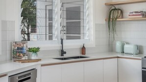 Moderni keittiö, jossa on valkoiset kaapit, ruostumattomasta teräksestä valmistettu pesuallas ja ikkunassa sälekaihtimet. Ikkunalaudalla on kasvi ja työtasolla kirja, jonka nimi on ”Wholesome”.