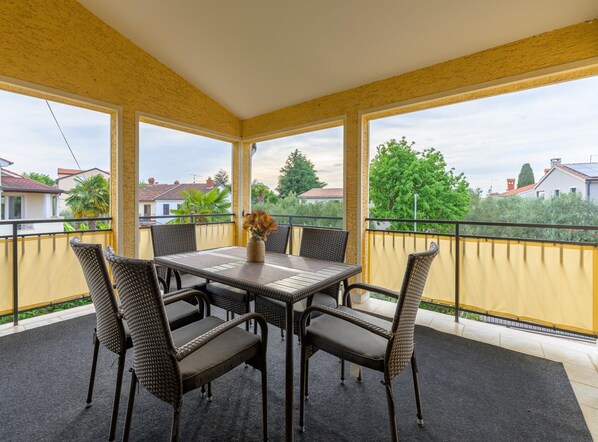 A covered balcony with a table and chairs, a vase with flowers, and a view of houses and trees.