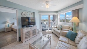 A spacious living room with a television, wicker furniture, a glass coffee table, and a balcony with a view.