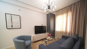 Ein modernes Wohnzimmer mit grauem Sofa, einem grauen Sessel, einem Flachbildfernseher auf einer Holzkommode, einem Glastisch und einem großen Fenster mit Vorhängen.