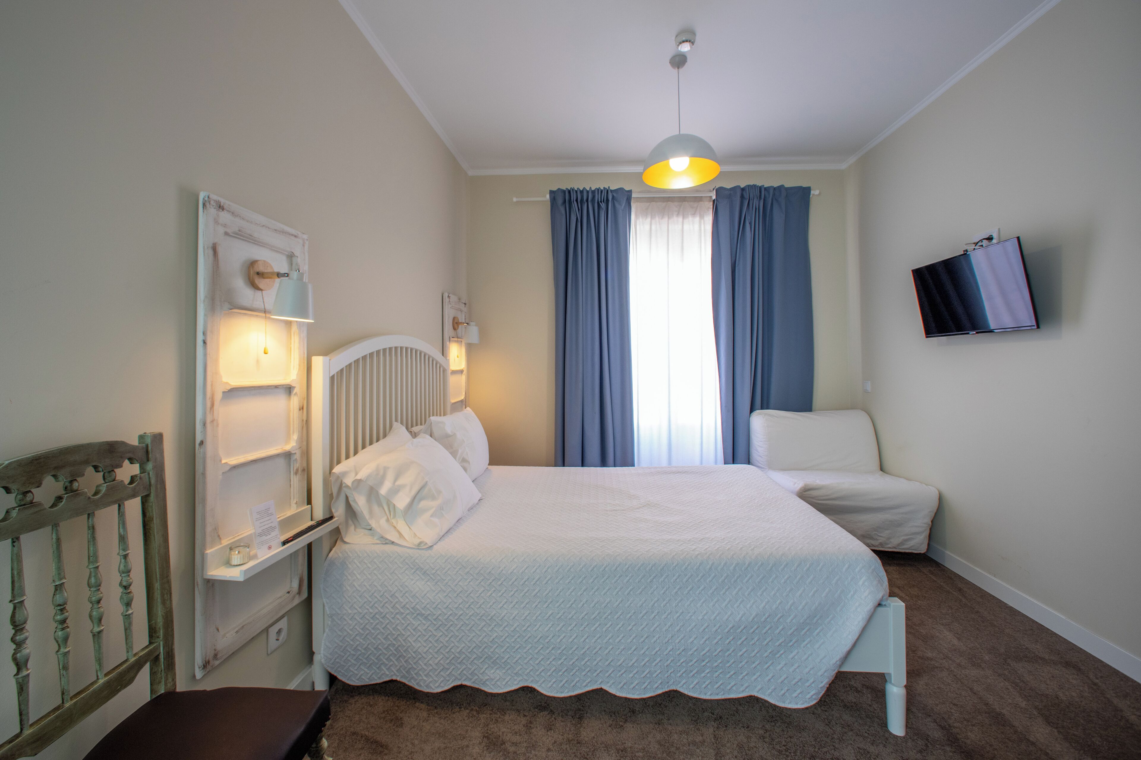 A bedroom with a bed, a nightstand, a dresser, a chair, a TV mounted on the wall, and a window with curtains.