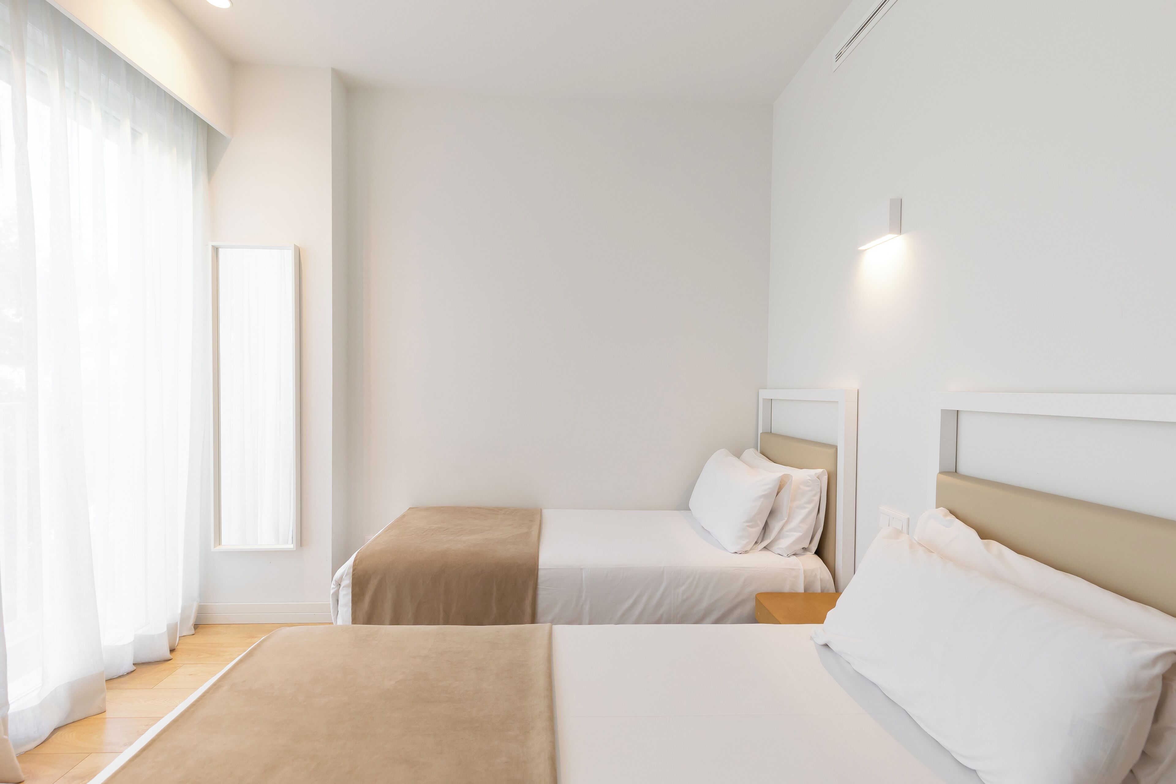 A hotel room with two beds, a large window with sheer curtains, and a wall-mounted light.