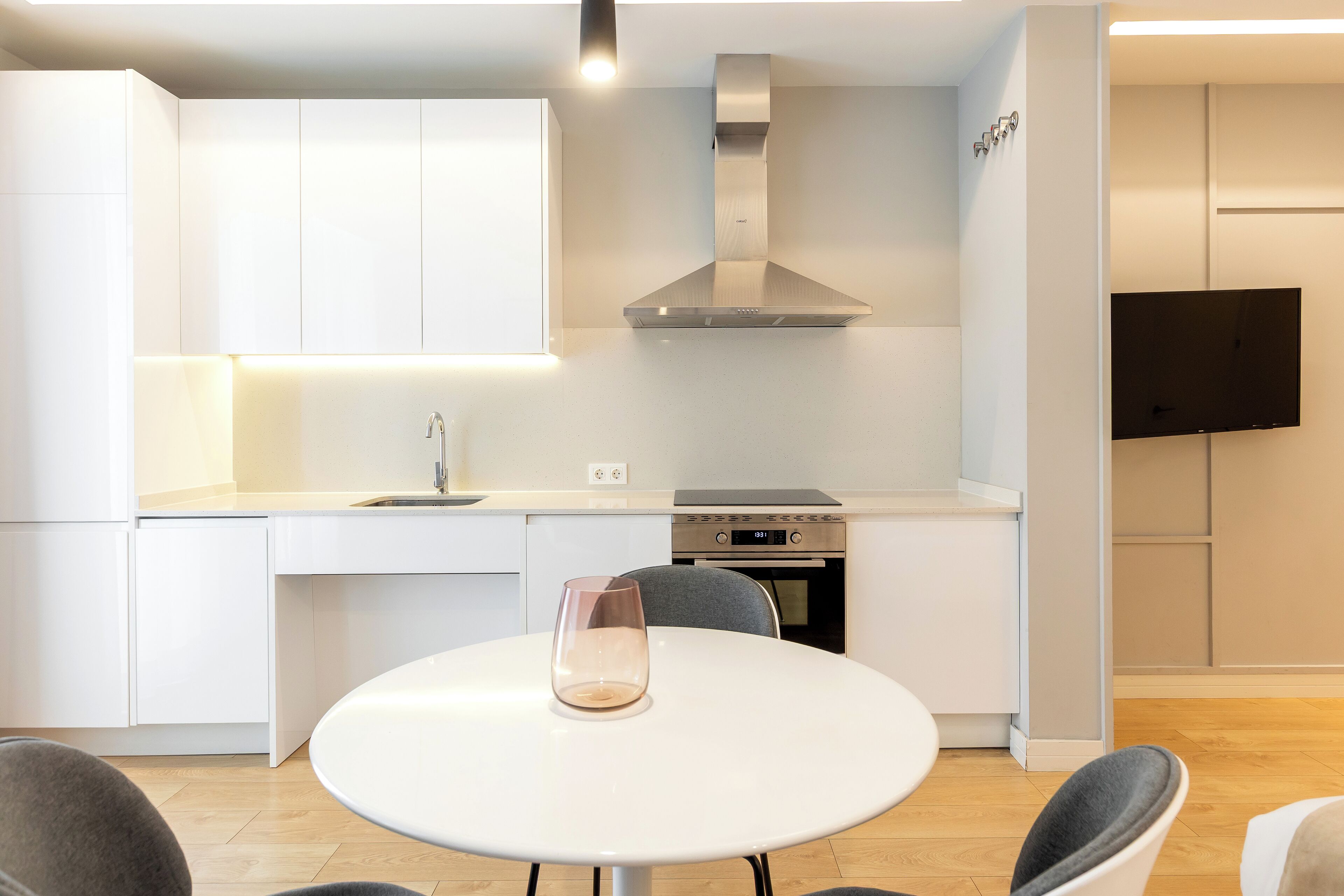 A modern kitchen with white cabinetry, a stainless steel range hood, a built-in oven, and a flat-screen TV mounted on the wall.