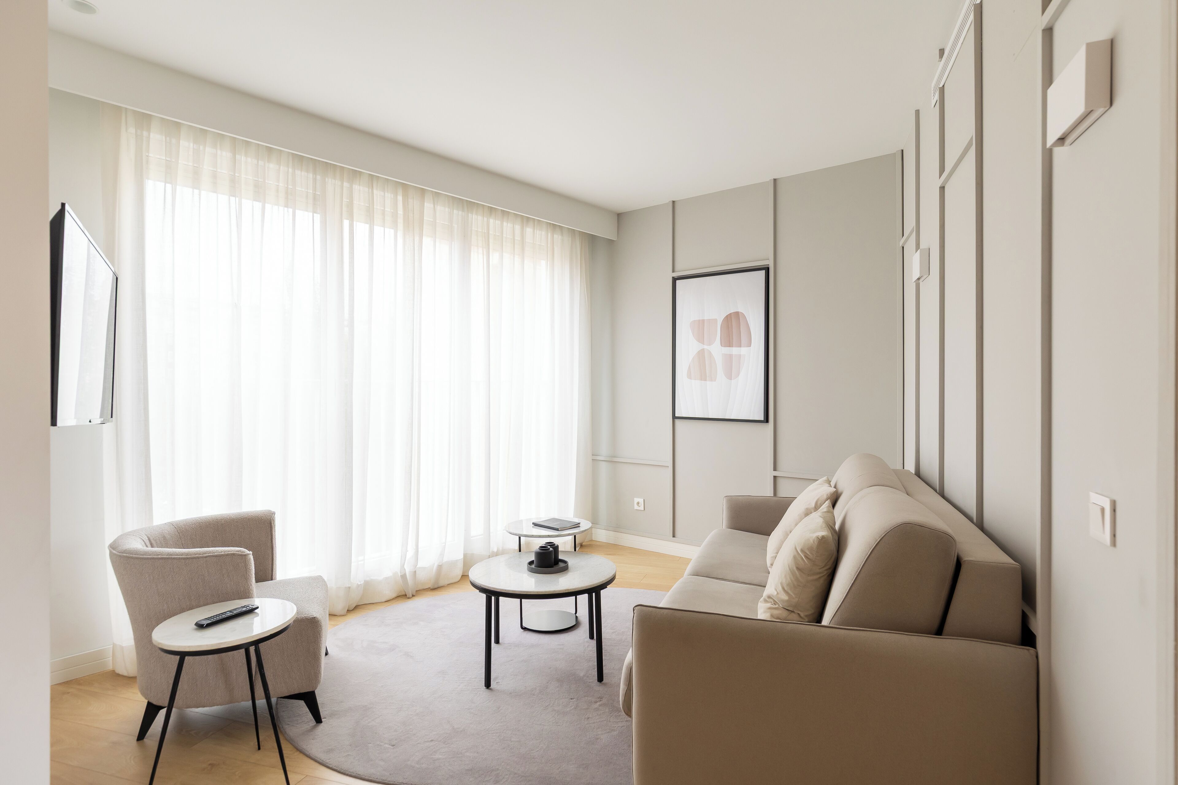 Una sala de estar moderna con sofá, sillón y mesa de centro, con un amplio ventanal con cortinas traslúcidas.