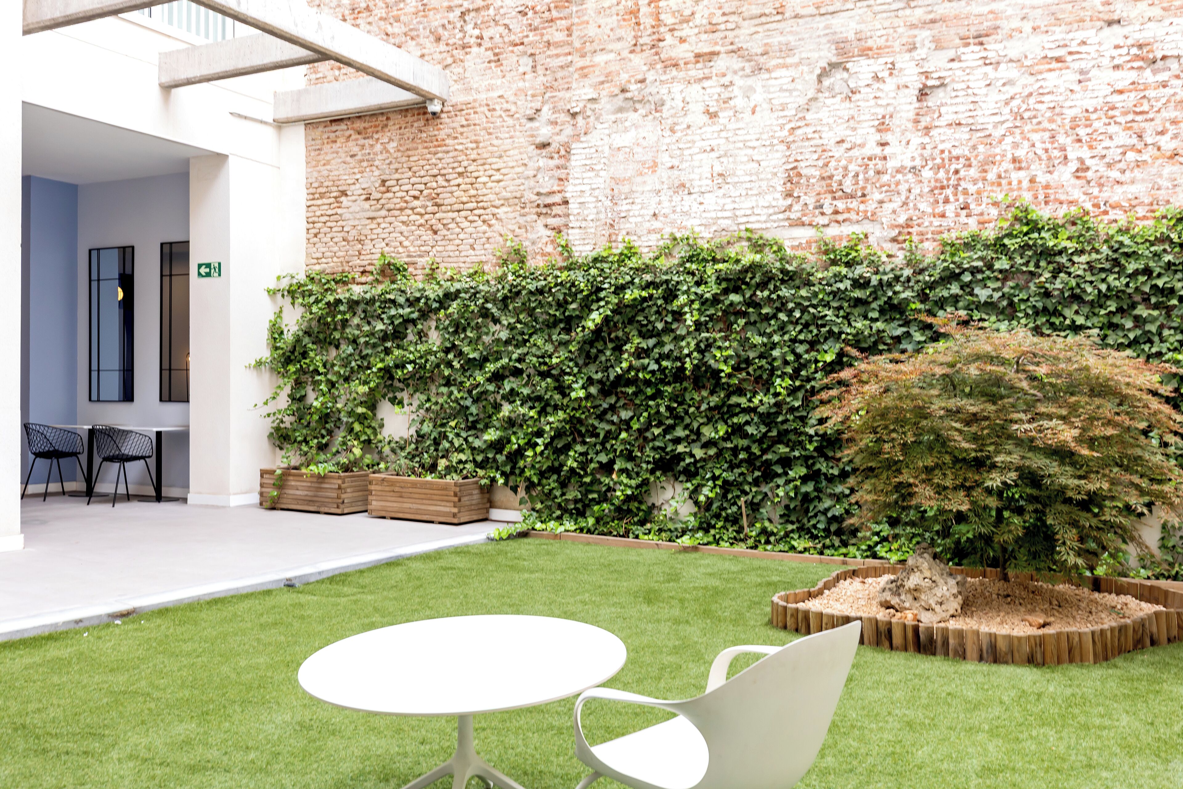 A courtyard with a brick wall, green hedge, and a small tree. There is a white table and two chairs in the foreground.