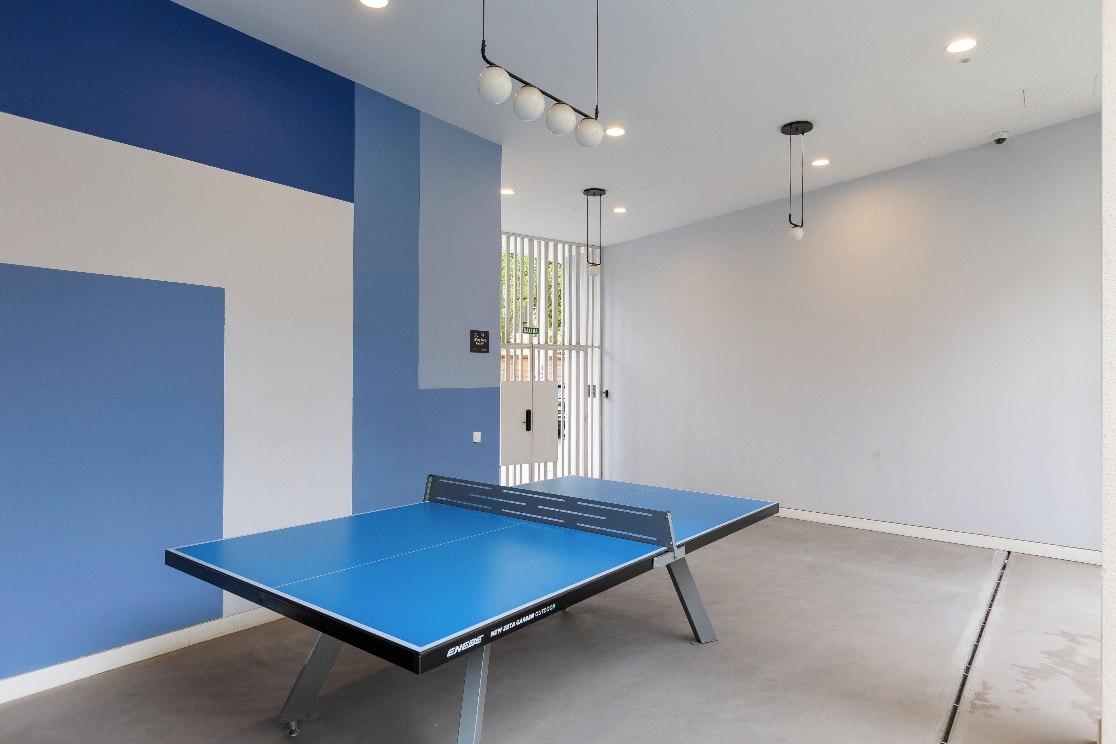 Un tavolo da ping pong in una stanza dalle pareti blu e bianche, con soffitto illuminato da faretti e una porta dotata di inferriata metallica.