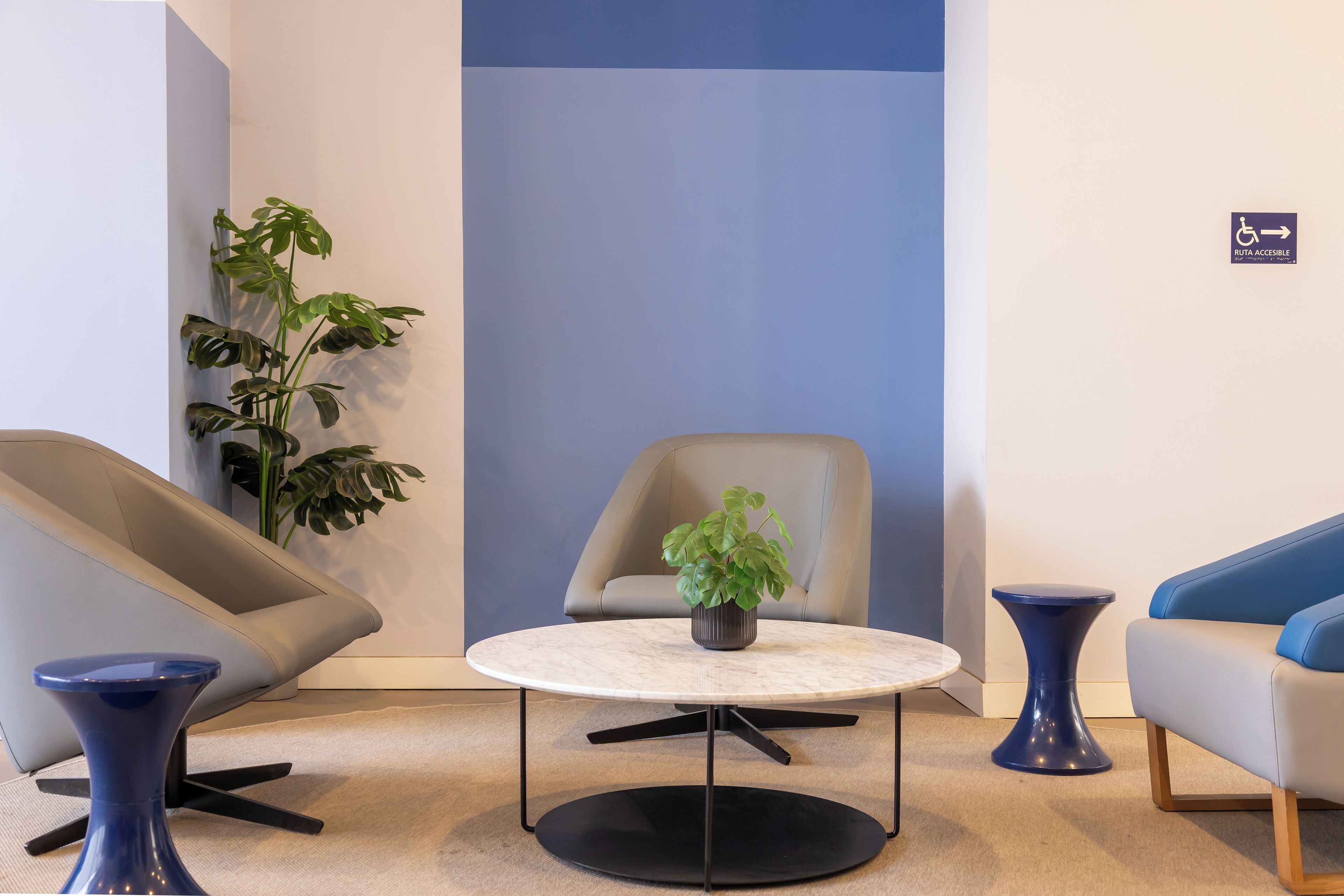 Uma área de estar moderna com uma mesa redonda, duas cadeiras, uma planta em vaso e uma placa azul indicando acessibilidade.