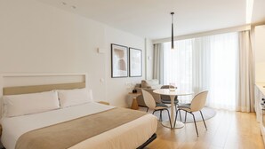 Ett hotellrum med en säng, ett litet runt bord med stolar, en soffa och ett stort fönster med gardiner.