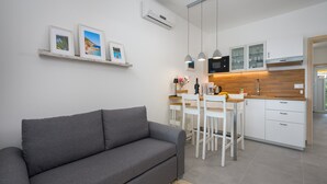 Eine moderne Küche mit weißen Schränken, einer Mikrowelle und einer Kaffeemaschine. Im Wohnbereich befindet sich ein graues Sofa und ein Esstisch mit weißen Stühlen.