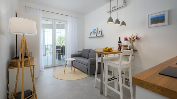 Ein modernes Wohnzimmer mit einer Couch, einem Esstisch, Stühlen, einer Lampe und Blick ins Freie durch eine Schiebetür.