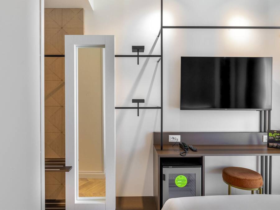 A hotel room with a bed, a desk with a chair, a television, a minibar, and a door leading to a bathroom.