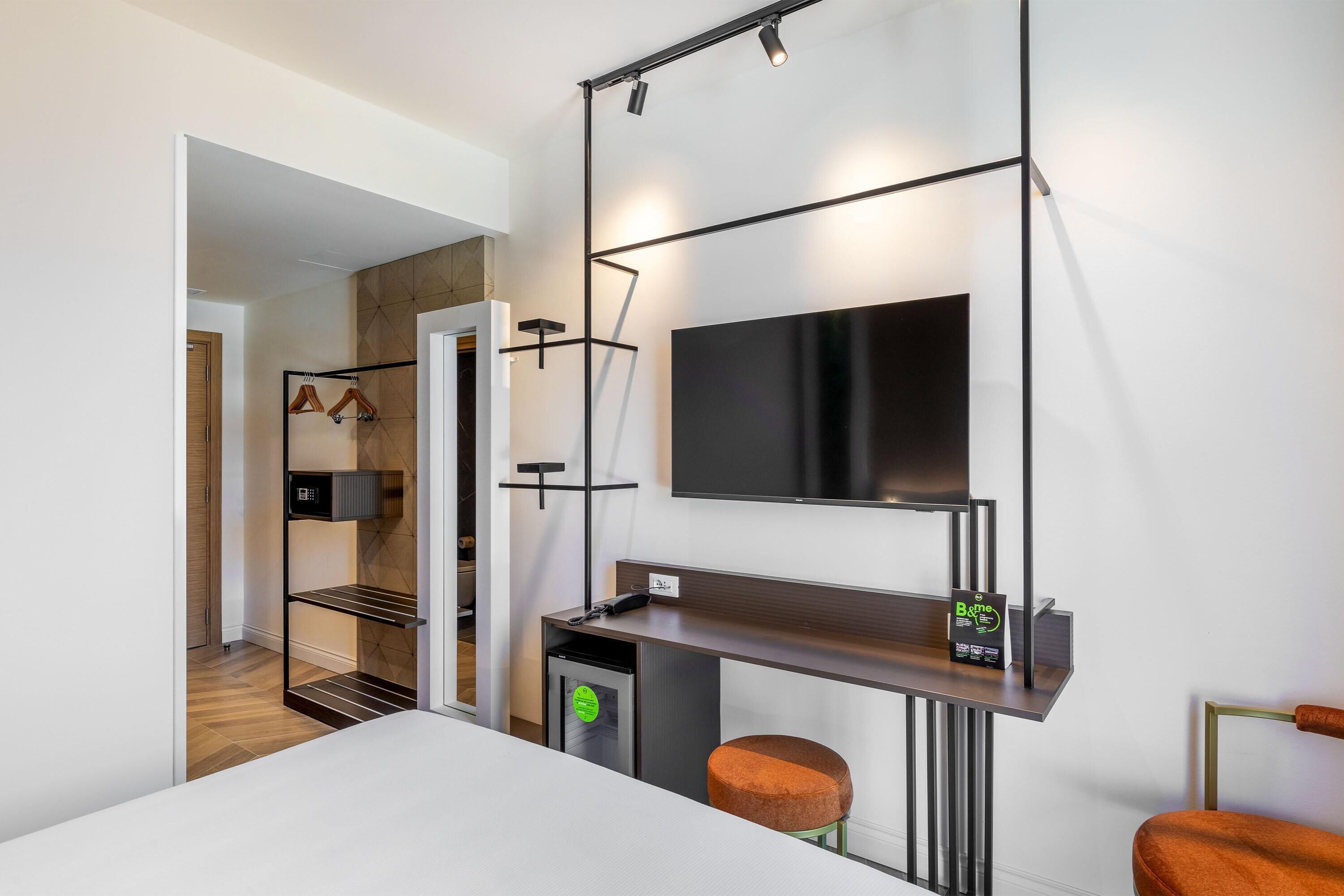 A modern hotel room with a flat-screen TV, a desk with a chair, a small fridge, and a wardrobe with hanging clothes.