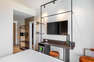 Una moderna camera d'hotel con televisione a schermo piatto, una scrivania con sedia, un piccolo frigorifero e un armadio con abiti appesi.