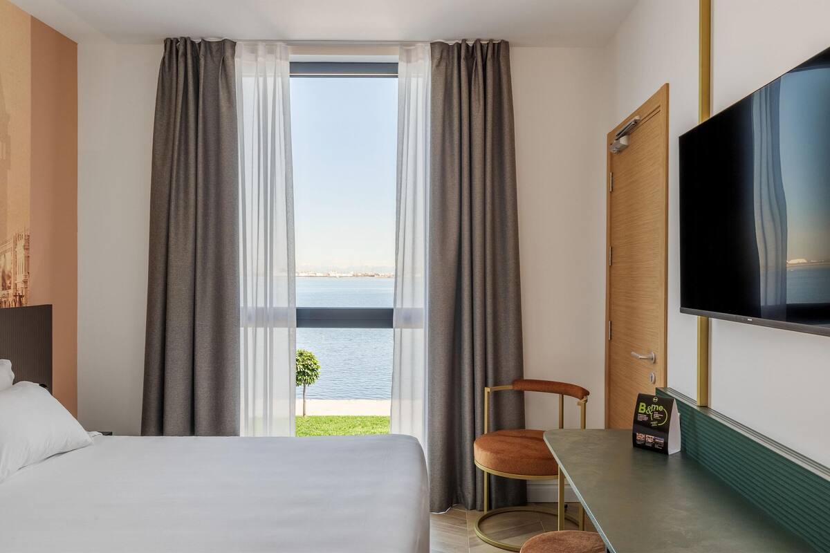 Hotelkamer met een bed, een televisie, een tafel met stoel, en uitzicht op een waterpartij door grote ramen.