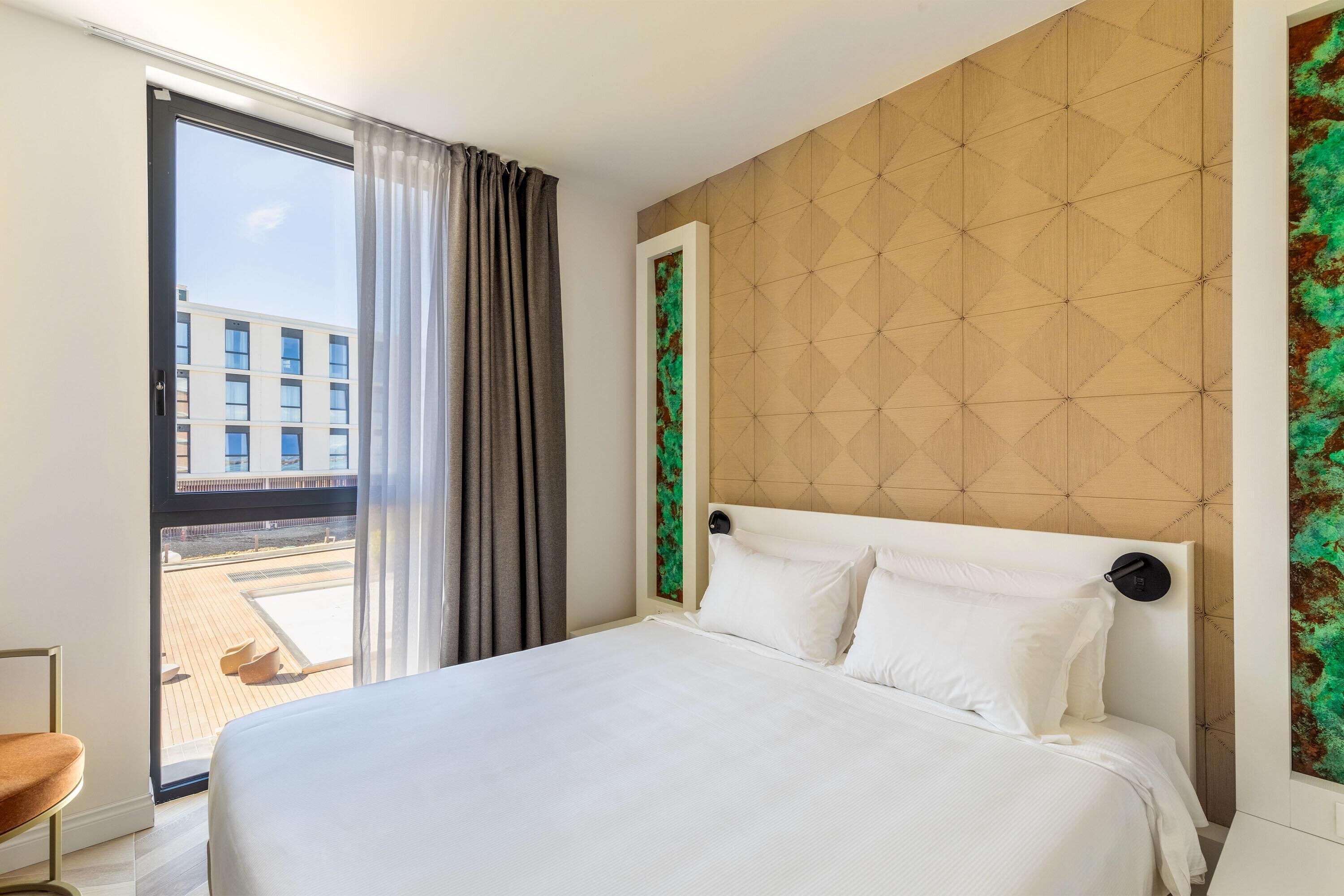 Hotelkamer met een bed, uitzicht naar buiten, een deur en een muur met een geometrisch patroon.