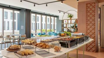 Ein Buffet mit verschiedenen Speisen, ein Sitzbereich mit Stühlen und Tischen sowie große Fenster mit Blick auf Gebäude.