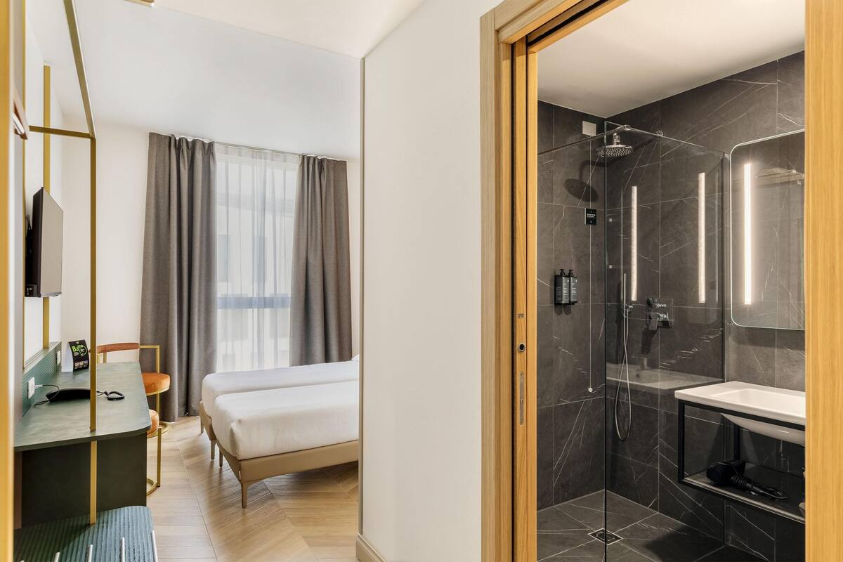 Hotelkamer met een bed, bureau met stoel, douche met glazen deur en een groot raam met gordijnen.